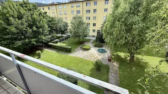 Blick vom Balkon auf den begrünten Innenhof mit Bäumen, Sträuchern und einem Wohngebäude im Hintergrund.
