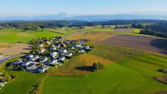 Luftaufnahme eines ländlichen Dorfes umgeben von grünen Feldern mit Bergen im Hintergrund.