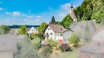 Ländliche Ansicht eines traditionellen Hauses mit Satteldach, umgeben von viel Grün und einer Kirche auf dem Hügel.