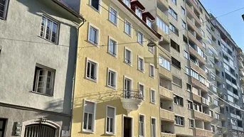 Gelbes Mehrfamilienhaus an einer sonnigen Straße mit parkenden Autos und angrenzenden Gebäuden.