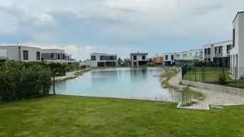 Wohngebiet mit einem zentralen See, umgeben von modernen Häusern und gepflegten Rasenflächen.