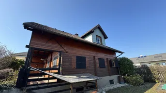 Rustikales Holzhaus mit Satteldach und Gartenbereich unter blauem Himmel an einem sonnigen Tag.