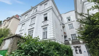 Historisches Gebäude mit weißer Fassade, vielen Fenstern und kleinen Balkonen, umgeben von Grün.