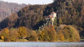 Historisches Gebäude auf einem Felsen über dem Fluss, umgeben von herbstlichen Wäldern.