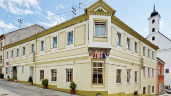 Historisches Gebäude in Gelb an einer Straßenecke mit traditioneller Architektur und Kirchturm im Hintergrund.