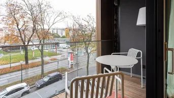 Balkon mit Holzboden, Sitzgelegenheiten und Blick auf die grüne Umgebung und Straße.