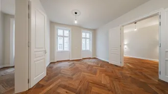 Geräumiges Zimmer mit klassischem Fischgrätparkett und zwei hohen Fenstern.