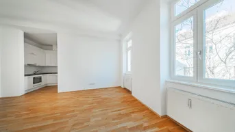 Helles Wohnzimmer mit Parkettboden und offener Küche, durch große Fenster lichtdurchflutet.