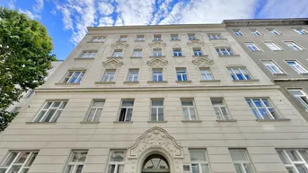 Historische Fassade eines Altbaus mit eleganten Verzierungen und großen Fenstern unter blauem Himmel.