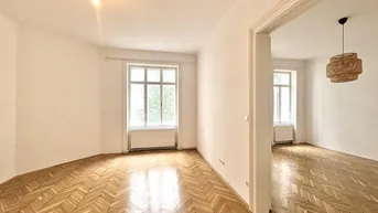 Heller Wohnraum mit glänzendem Fischgrätparkett und hohen Decken, der in einen weiteren Raum führt.