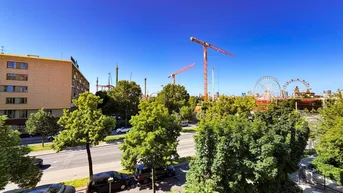 Blick auf die Stadt mit Bäumen, Straße und dem Prater im Hintergrund bei blauem Himmel.