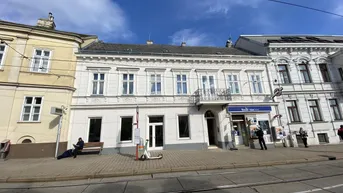 Historisches Gebäude mit klassischer Fassade, kleinem Balkon und Geschäften im Erdgeschoss an einer belebten Straße.