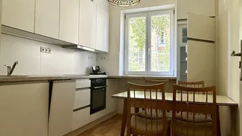 Helle Küche mit weißen Schränken, Holzboden und einem Esstisch am Fenster.