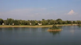 Weiter Blick über einen ruhigen See mit bewaldetem Ufer und kleinen Häusern im Hintergrund.