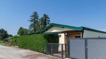 Einfamilienhaus mit gelber Fassade, grüner Hecke und modernem Eingangstor an einem sonnigen Tag.