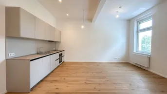 Helle, moderne Küche mit hellgrauen Schränken, Holzboden und großem Fenster, Teil eines offenen Wohnbereichs.