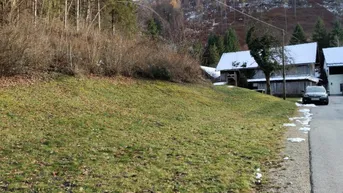 Blick auf eine grüne Wiese mit leichtem Schneefall, umgeben von bewaldeten Hügeln und ländlichen Gebäuden.