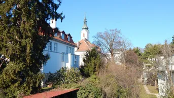 Blick auf ein historisches Gebäude mit Kirchturm und umliegender grüner Landschaft unter blauem Himmel.