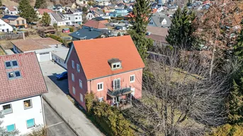 Luftaufnahme eines Einfamilienhauses mit rotem Dach und umliegender Wohnsiedlung an einem sonnigen Tag.