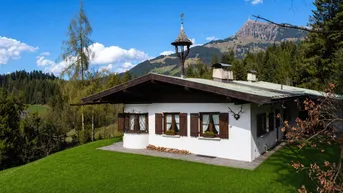 Weißes Haus im alpenländischen Stil mit Holzelementen, umgeben von grüner Wiese und Bergen.