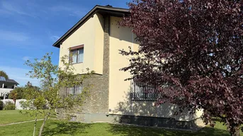 Seitenansicht des Hauses mit heller Fassade, Steinelementen und gepflegtem Vorgarten unter blauem Himmel.