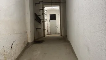 Langer, schmaler Kellerflur mit weiß gestrichenen Wänden und Betonboden, der zu einer Tür führt.
