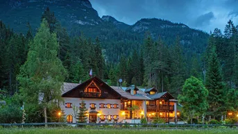Traditionelles Alpenhaus bei Dämmerung, umgeben von Wald und Bergen, mit warm beleuchteten Fenstern.