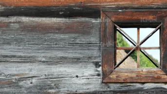 Alte, verwitterte Holzwand mit einem kleinen, rustikalen Fenster und Blick ins Grüne.