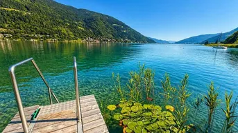 Klarer Bergsee mit Holzsteg und Badeleiter, umgeben von grünen Bergen unter blauem Himmel.