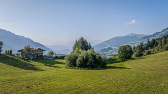 Panoramablick auf ein traditionelles Haus in einer grünen Berglandschaft unter blauem Himmel.
