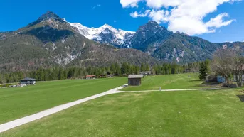 Weitläufige grüne Wiese mit majestätischen Bergen im Hintergrund und vereinzelten Häusern.