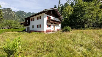 Traditionelles Haus im alpenländischen Stil mit roten Fensterläden und Holzbalkonen, umgeben von Natur und Bergen.