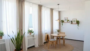 Helles Wohnzimmer mit Laminatboden, großen Fenstern und einem kleinen Essbereich, dekoriert mit Pflanzen.
