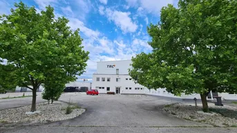 Weißes Geschäftsgebäude mit Firmenlogo, umgeben von Bäumen und einem Parkplatz unter blauem Himmel.