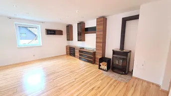 Geräumiges Wohnzimmer mit Laminatboden, einem gemütlichen Holzofen und modernen dunklen Möbeln.