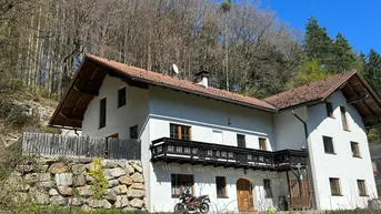 Traditionelles Haus mit weißer Fassade und Holzbalkon, umgeben von bewaldeten Hügeln.