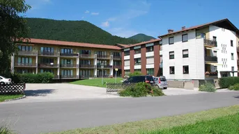 Mehrfamilienhaus mit mehreren Balkonen und Parkplätzen, umgeben von grüner Landschaft und Bergen.