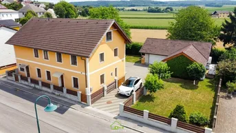 Haus mit gelber Fassade, braunem Dach und gepflasterter Einfahrt, umgeben von Grünflächen.