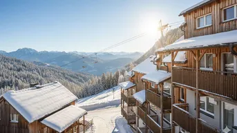 Holzchalets im Schnee mit Skilift und sonnigem Bergblick, umgeben von verschneiten Wäldern.
