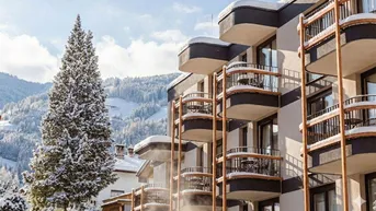 Modernes Apartmentgebäude mit Balkonen und verschneiter Berglandschaft im Hintergrund unter blauem Himmel.