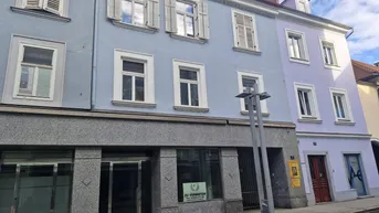 Ansicht der Gebäudefassade mit Geschäften im Erdgeschoss und Wohnungen darüber, mit blauen und lila Farbtönen.