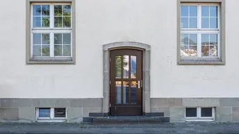 Weiße Fassade eines Gebäudes mit brauner Eingangstür und symmetrischen Fenstern im Erdgeschoss und Obergeschoss.