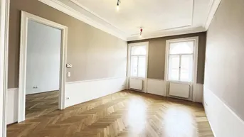 Helles Zimmer mit Fischgrätparkett, hohen Decken und Stuckverzierungen an den Wänden.