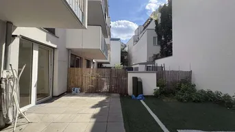 Große gepflasterte Terrasse mit angrenzendem Kunstrasenbereich und Blick auf die moderne Wohnanlage.