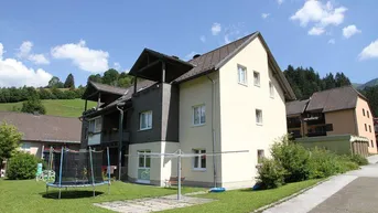Mehrfamilienhaus mit gepflegtem Garten, Trampolin und Balkonen in ländlicher Umgebung unter blauem Himmel.