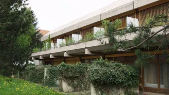Gebäude mit mehreren Balkonen und Holzjalousien, umgeben von üppigem Grün und einem Rasen.