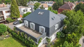 Luftaufnahme eines Hauses mit Mansarddach, großer Terrasse und gepflegtem Garten in grüner Umgebung.