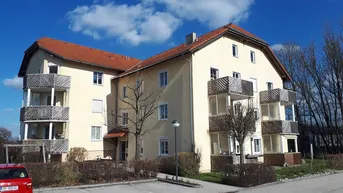 Mehrfamilienhaus mit beiger Fassade, roten Ziegeldächern und mehreren Balkonen in einer grünen Umgebung.