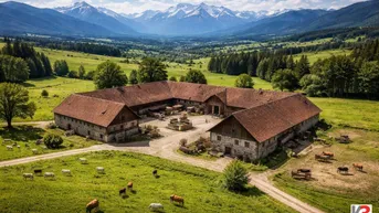 Luftaufnahme eines weitläufigen Anwesens mit mehreren Gebäuden, umgeben von grünen Wiesen und majestätischen Bergen im Hintergrund.