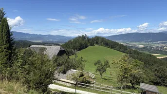 Ländliche Landschaft mit einem Haus, grünen Hügeln und Bergen im Hintergrund unter blauem Himmel.